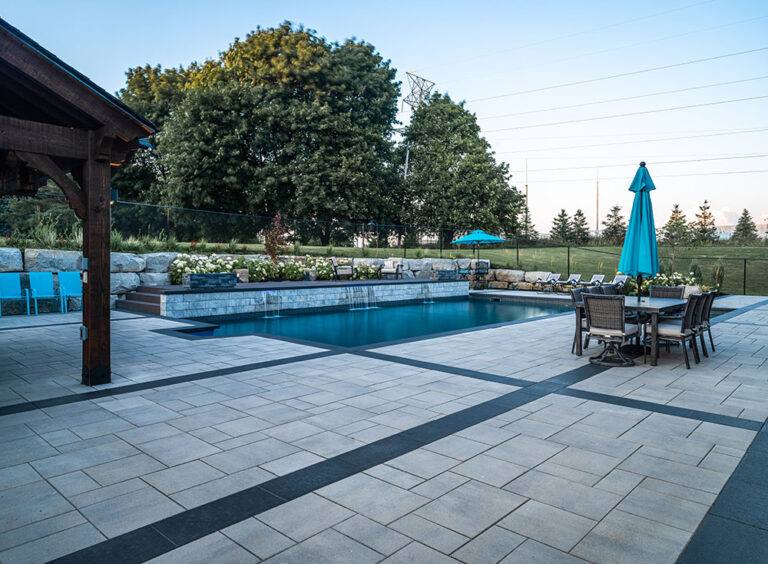 Wide-angle view of a modern backyard pool and outdoor dining area with trees.