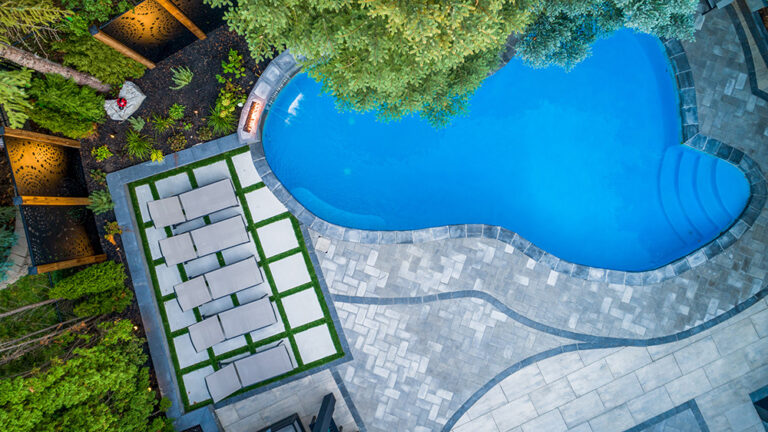 Elegant backyard pool design with surrounding stone patio and lush green landscaping.
